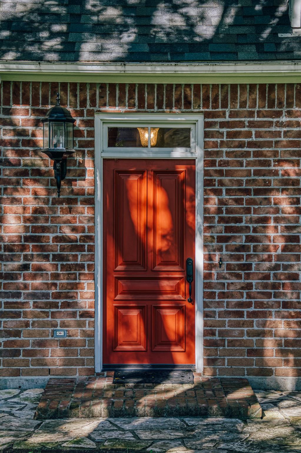 Red paneled front door in a brick facade, with a black lantern and stone walkway.