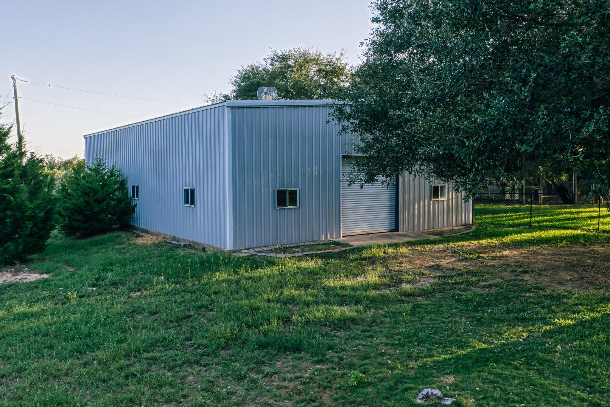 Gray metal barn with a roll-up door and small windows beside a grassy, tree-lined yard.