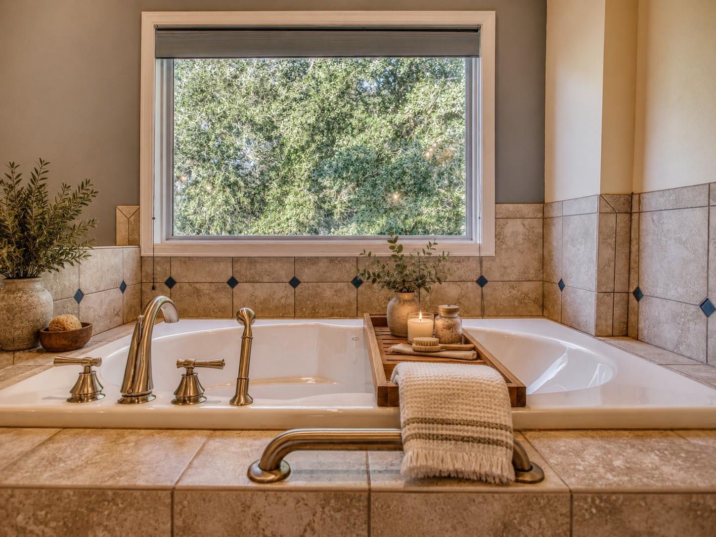 Soaking tub with brass fixtures, beige stone tile, and a large window overlooking greenery.