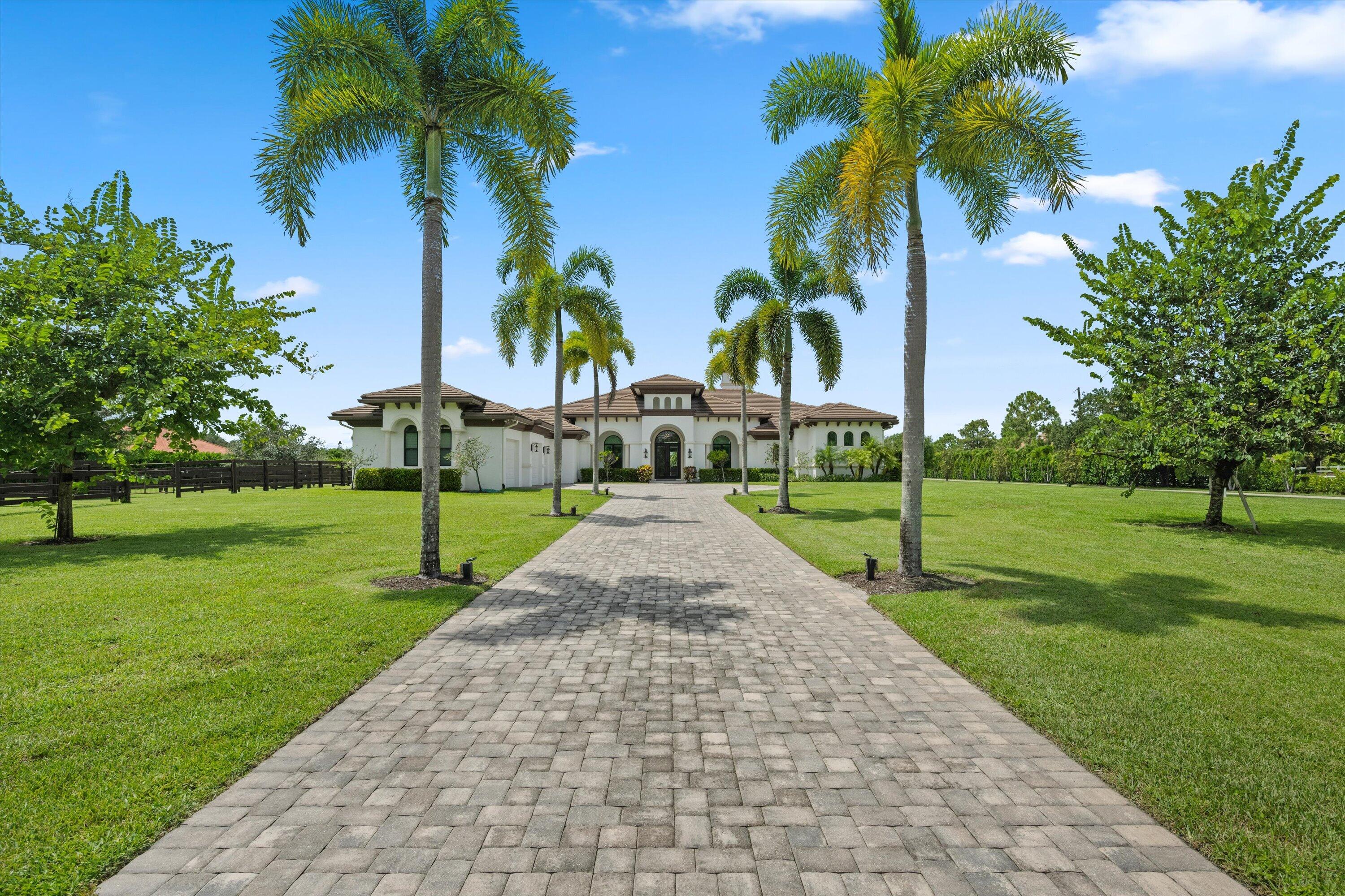 White stucco home beyond a broad paver driveway lined with tall palm trees.