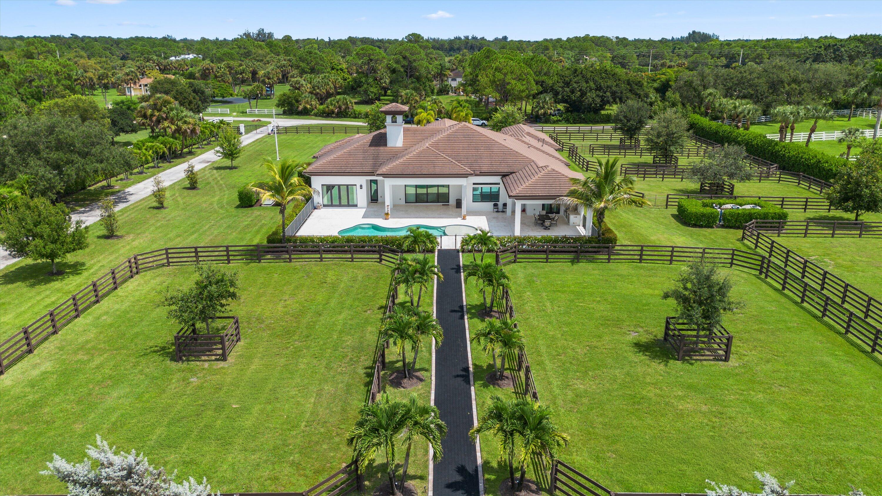 Aerial view of a home with a pool, palm-lined walkway, and fenced paddocks.