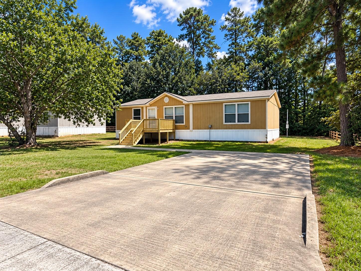 Tan single-story home with a raised front porch and wide concrete driveway, surrounded by mature trees.