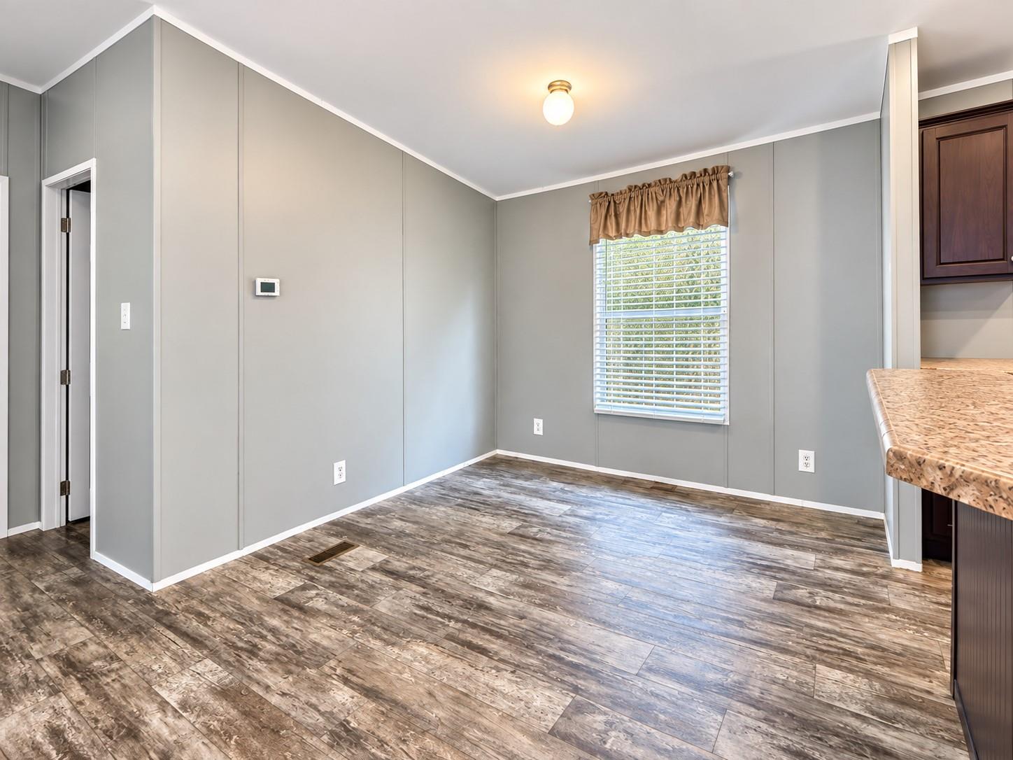 Empty dining area with gray paneled walls, distressed wood-look flooring, and a window with blinds.