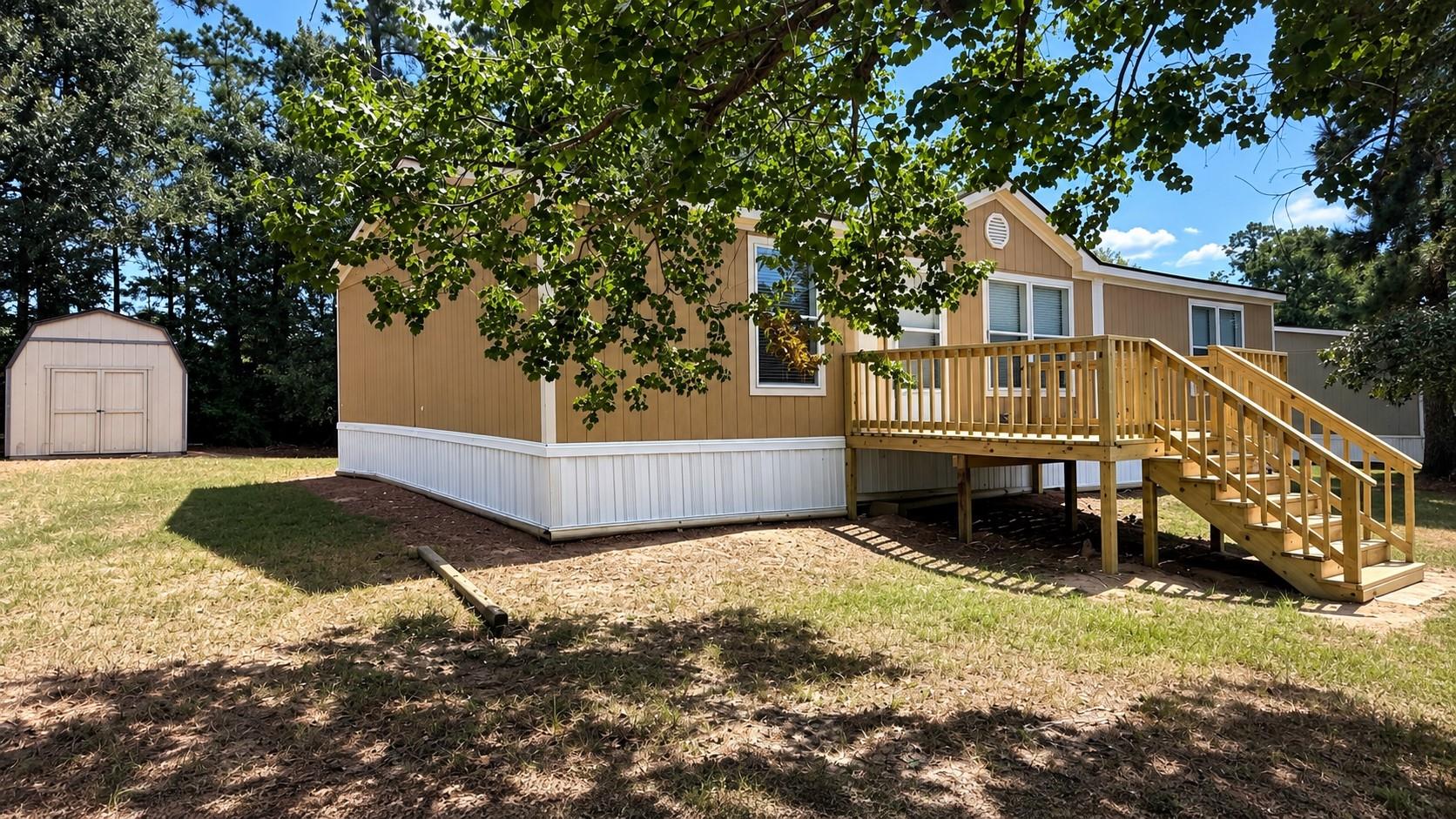 Tan manufactured home with a raised wooden deck and stairs, beside a storage shed in a tree-shaded yard.
