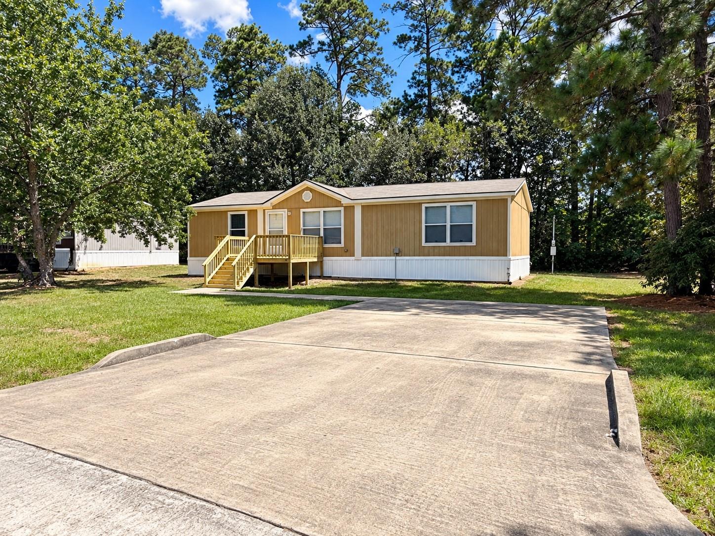Tan manufactured home with a raised wooden porch and broad concrete driveway beneath tall trees.