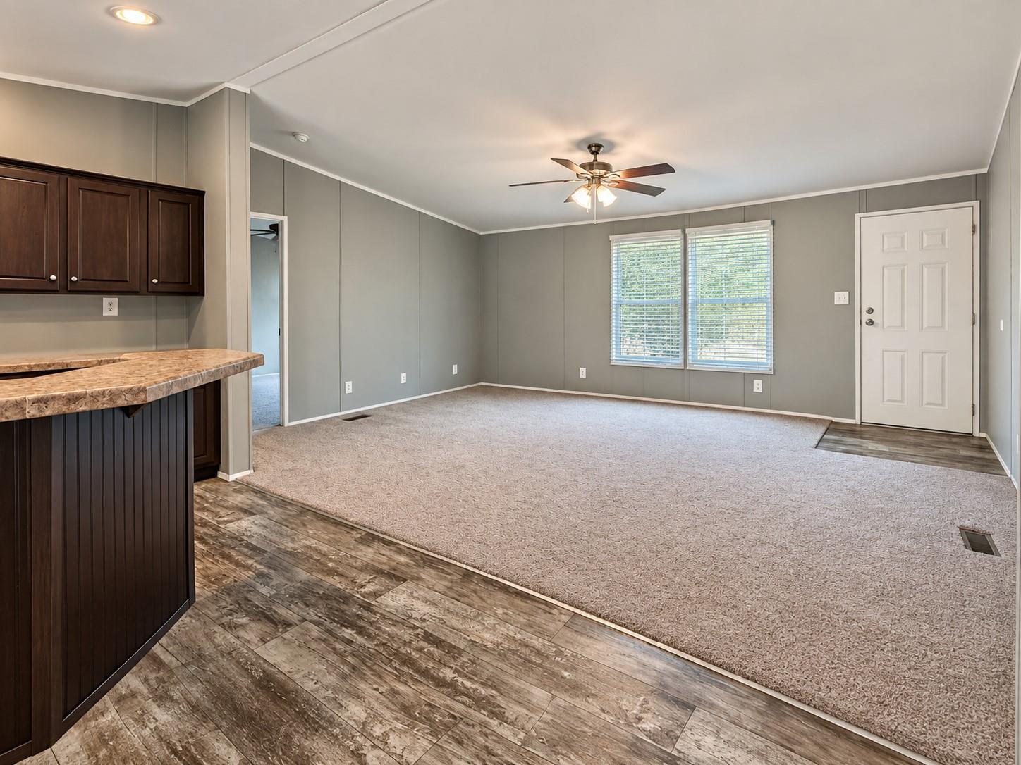 Vacant living room with gray walls, ceiling fan, two blinded windows, and a breakfast bar.