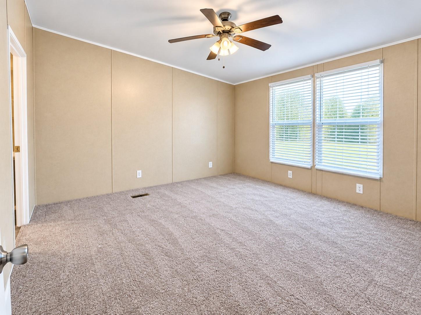 Empty bedroom with beige paneled walls, patterned carpet, two windows, and a ceiling fan.
