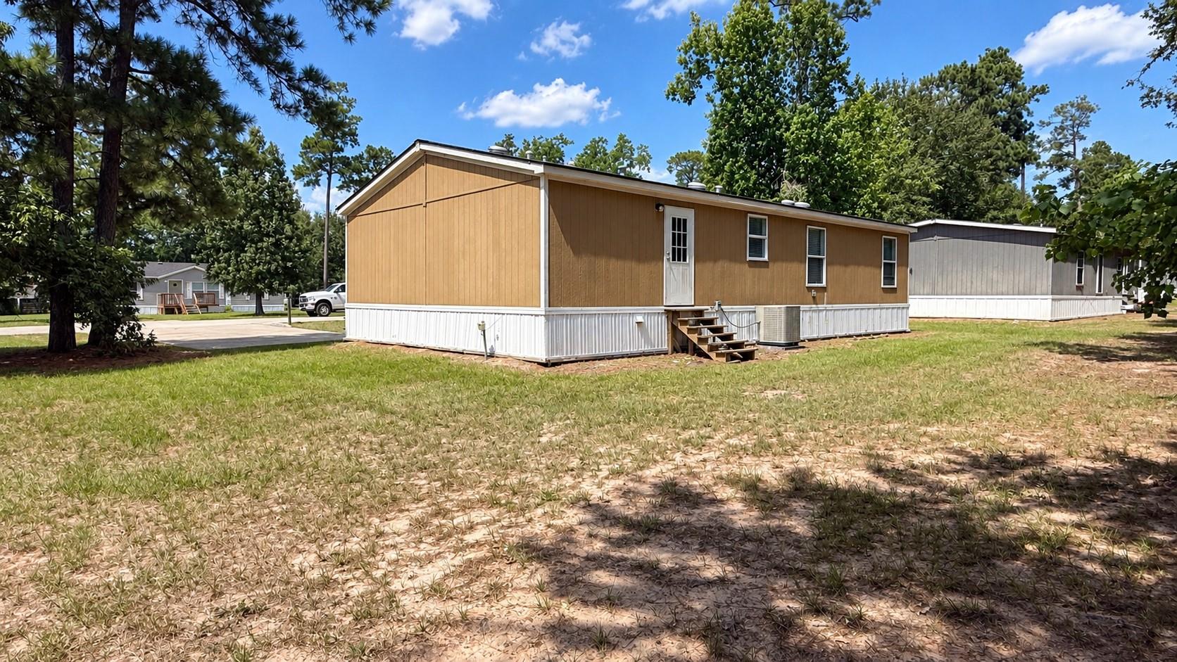 Tan manufactured home with white skirting and wooden steps on a tree-lined lot.