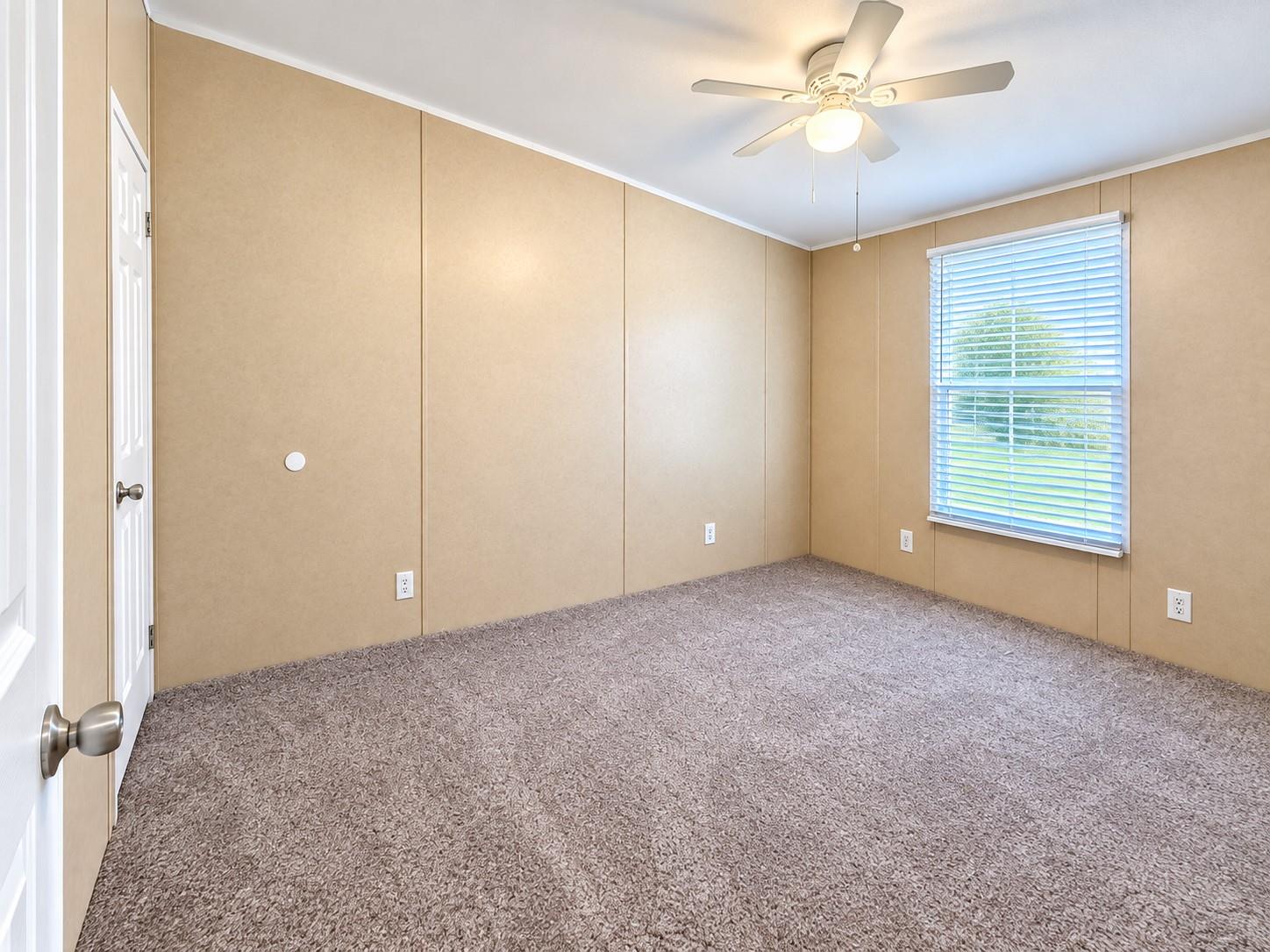 Empty bedroom with tan paneled walls, gray carpet, ceiling fan, and a window with blinds.