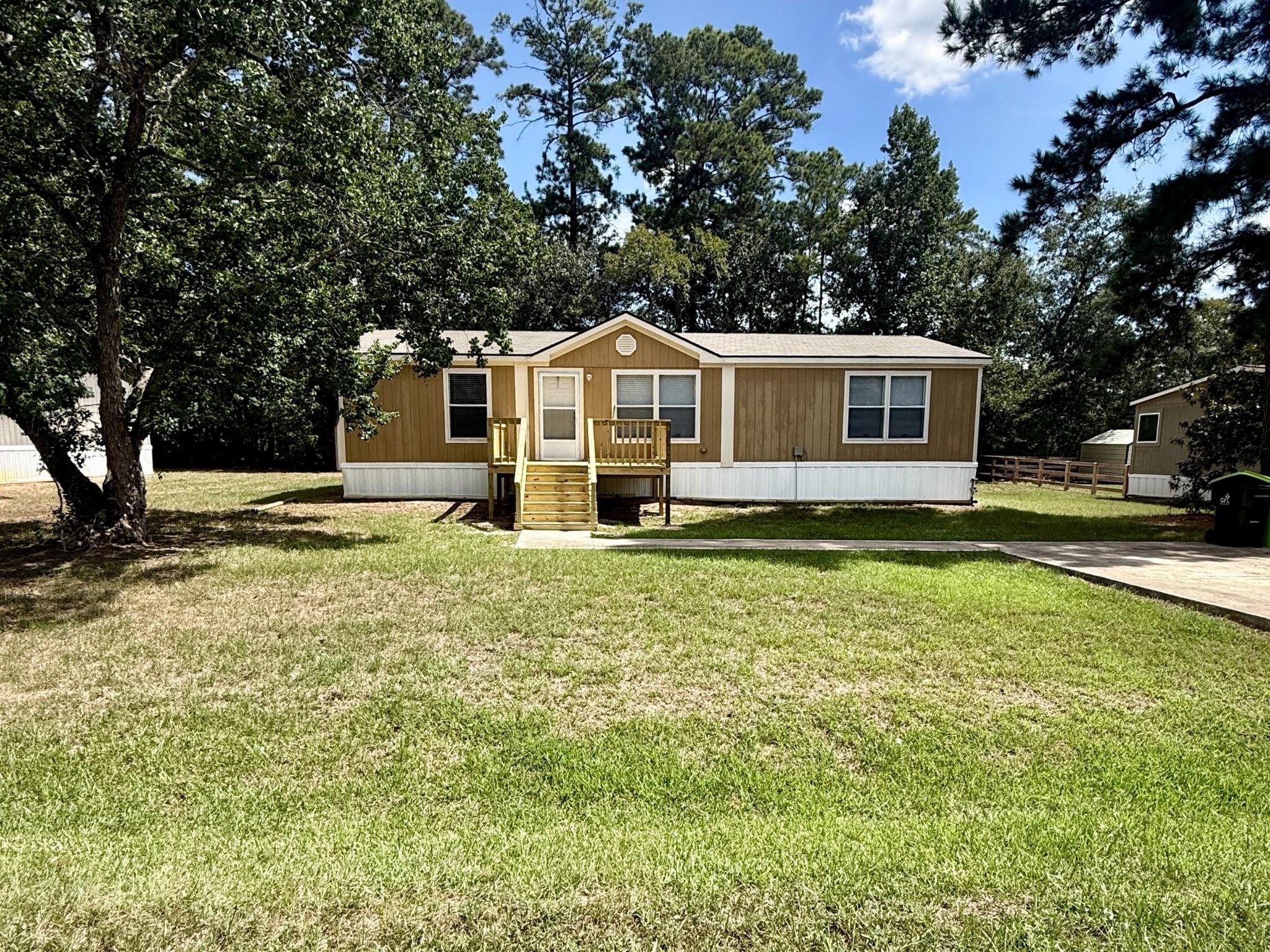Tan manufactured home with a small wooden front porch, broad lawn, and mature trees.