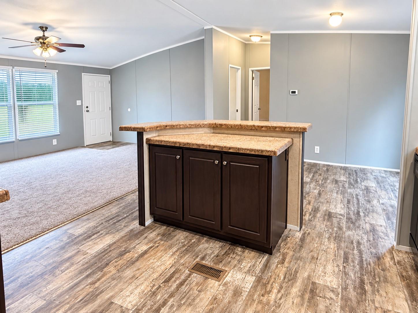 Open living area with a dark wood island, patterned countertop, ceiling fan, and windows with blinds.