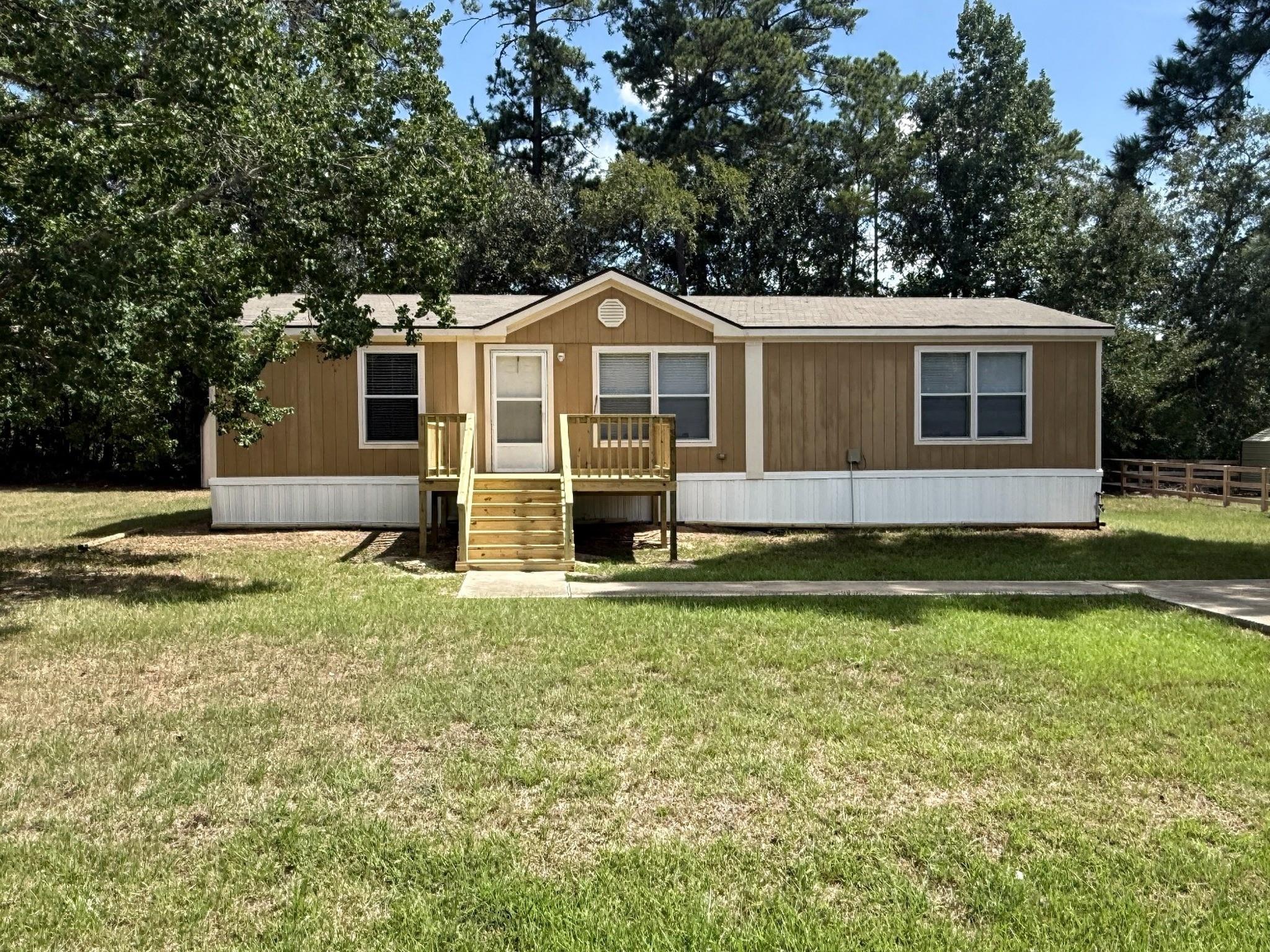 Brown single-story manufactured home with a raised wooden porch and steps, surrounded by a grassy lawn and mature trees.