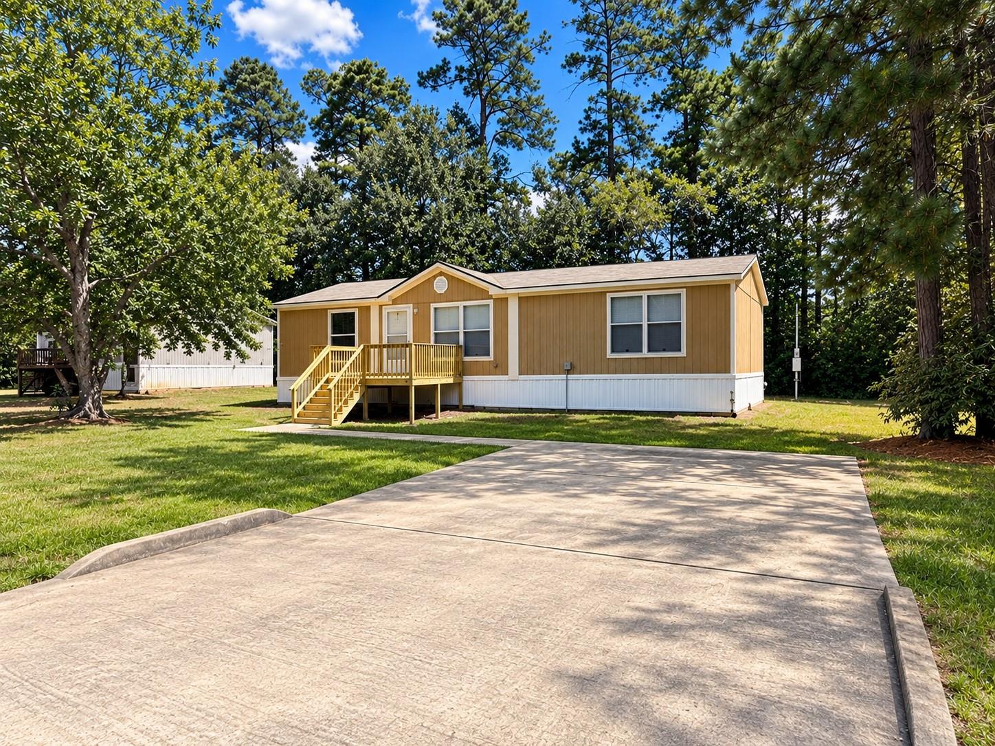 Tan single-story home with a raised porch and broad concrete driveway, surrounded by mature trees.