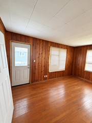 Empty wood-paneled room with polished hardwood floors, two windows, and a gray exterior door.