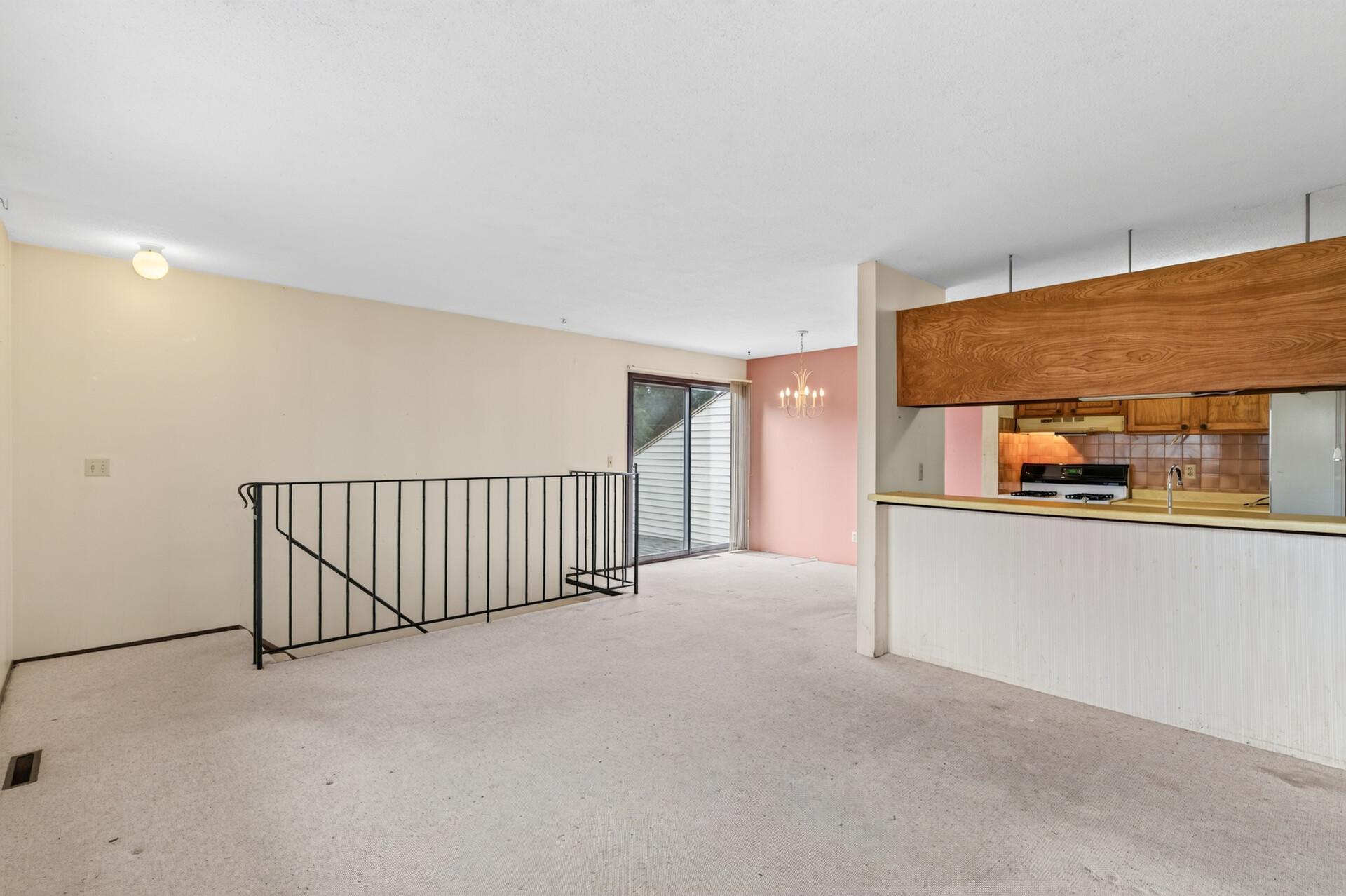 Carpeted living area with a stairwell railing, sliding glass door, and wood-cabinet kitchen pass-through.