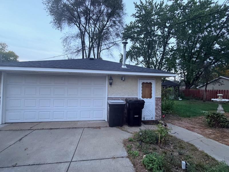 Garage exterior with a wide white paneled door, concrete driveway, and mature trees behind it.