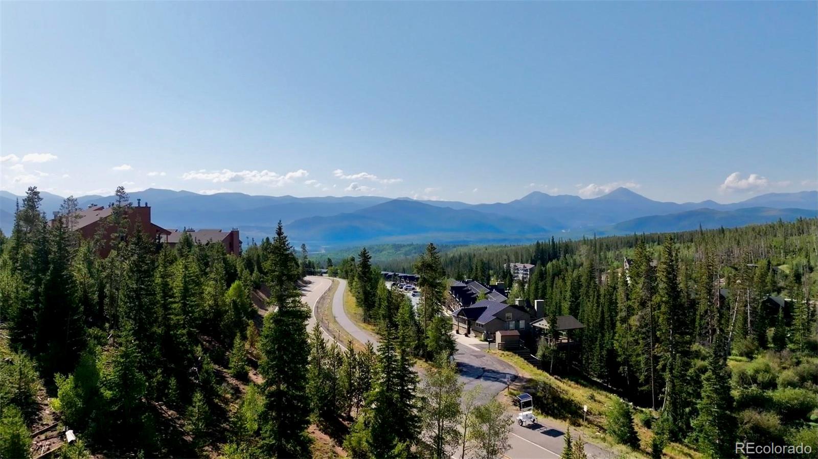 Aerial view of hillside homes among evergreens, with a winding road and distant mountain ranges.