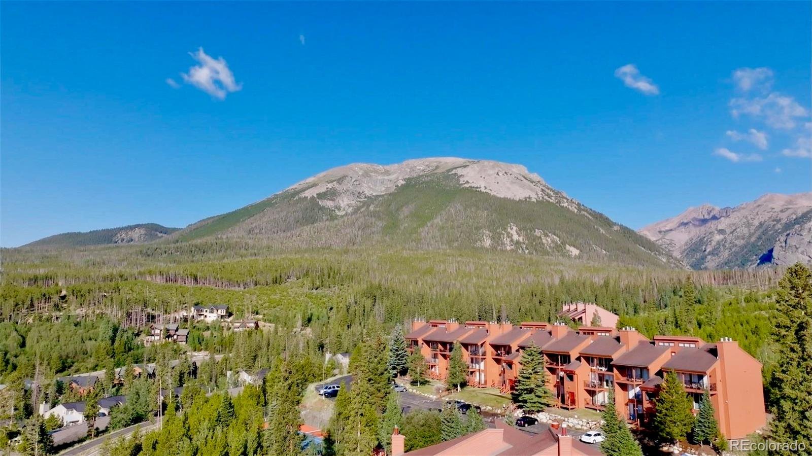Mountain condo complex beneath a broad rocky peak, surrounded by forest.