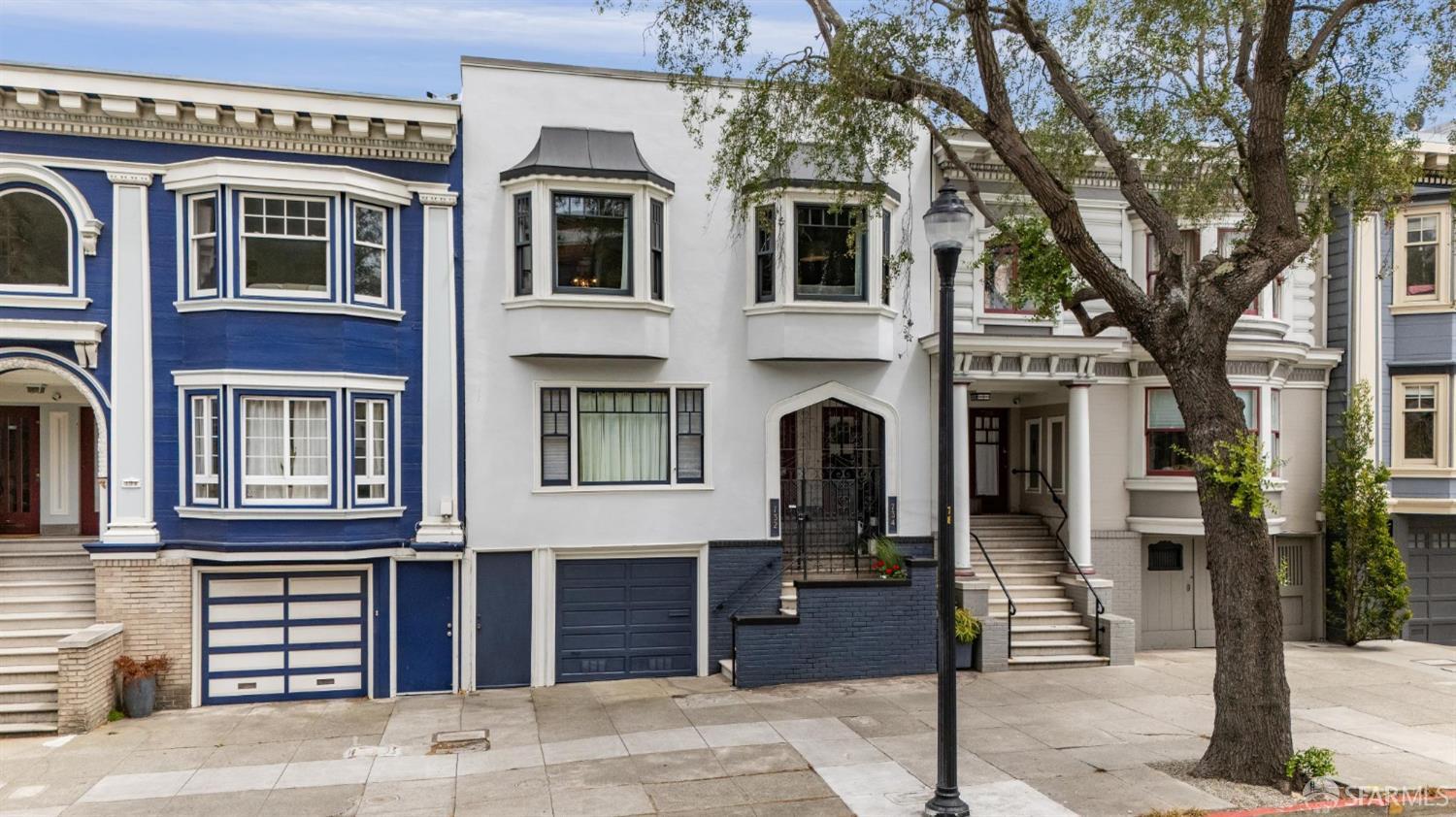 Attached San Francisco homes with bay windows, a dark blue garage, and a large sidewalk tree.