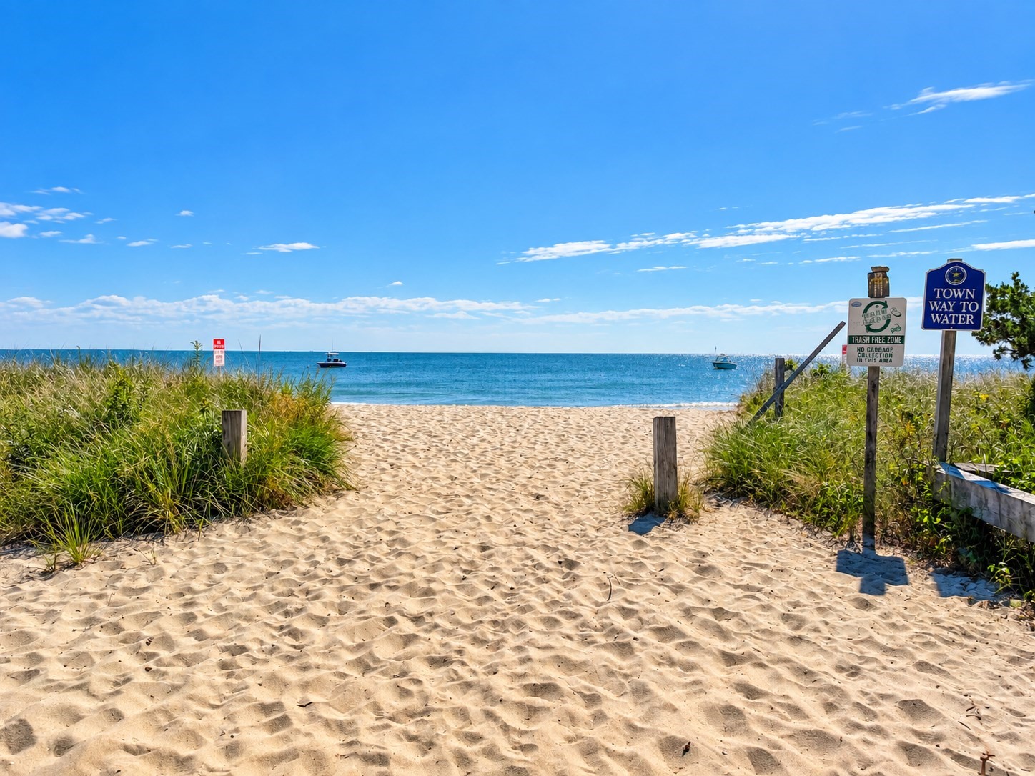Sandy beach access path to blue water, bordered by dune grass and marked with town signs.