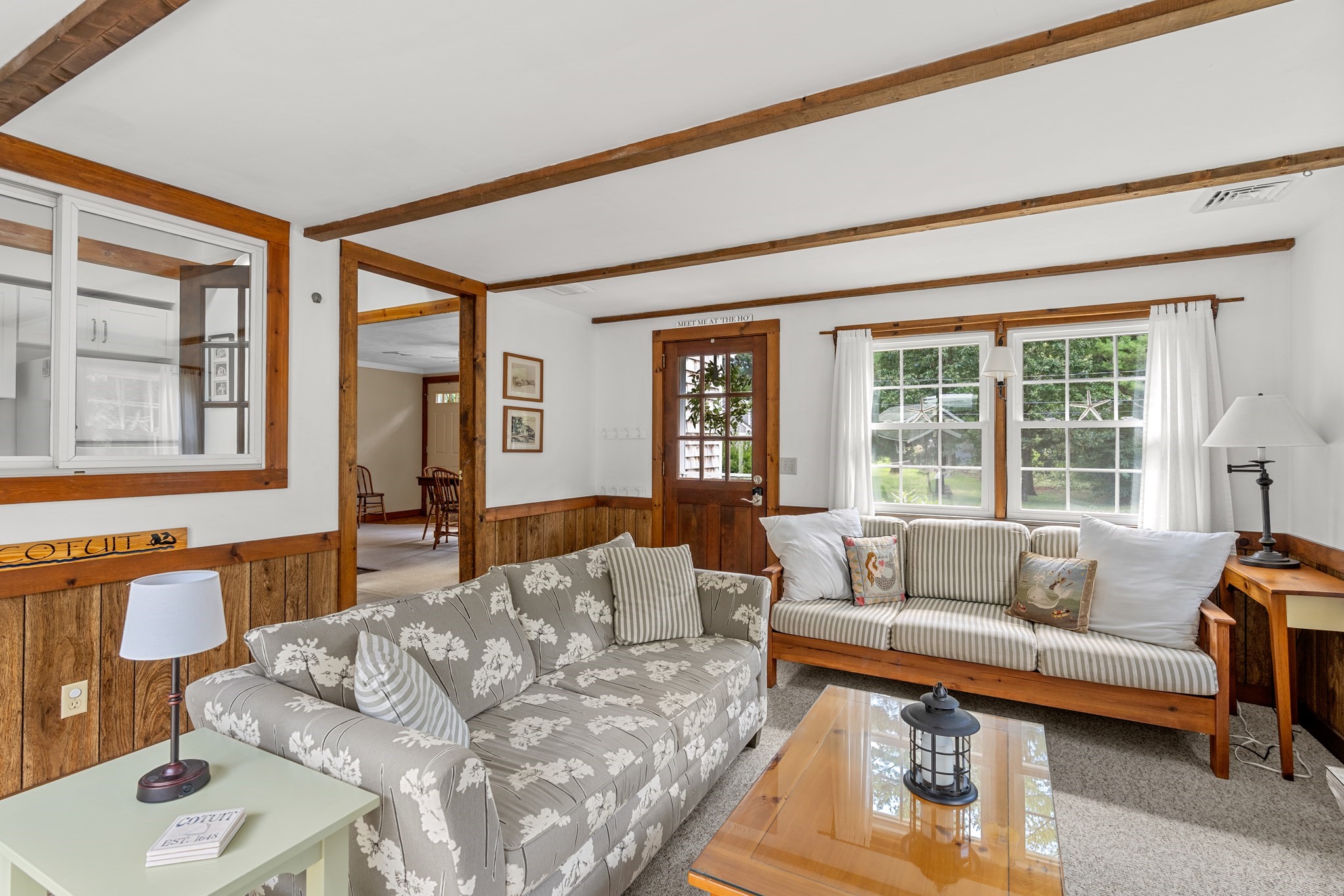 Living room with exposed wood beams, paneled walls, patterned sofas, and large curtained windows.