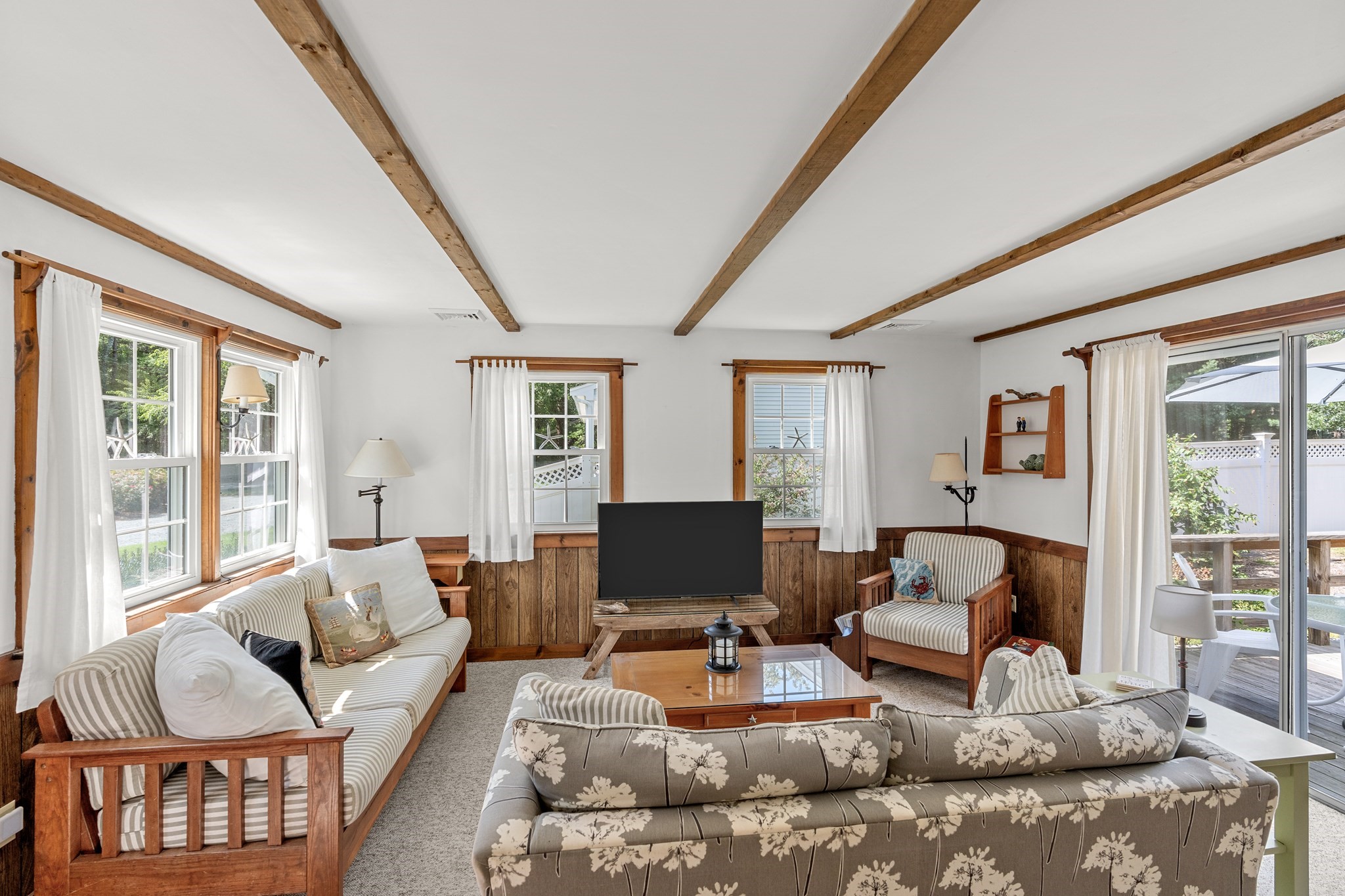 Living room with exposed wood beams, striped seating, wood paneling, and large windows opening onto a deck.