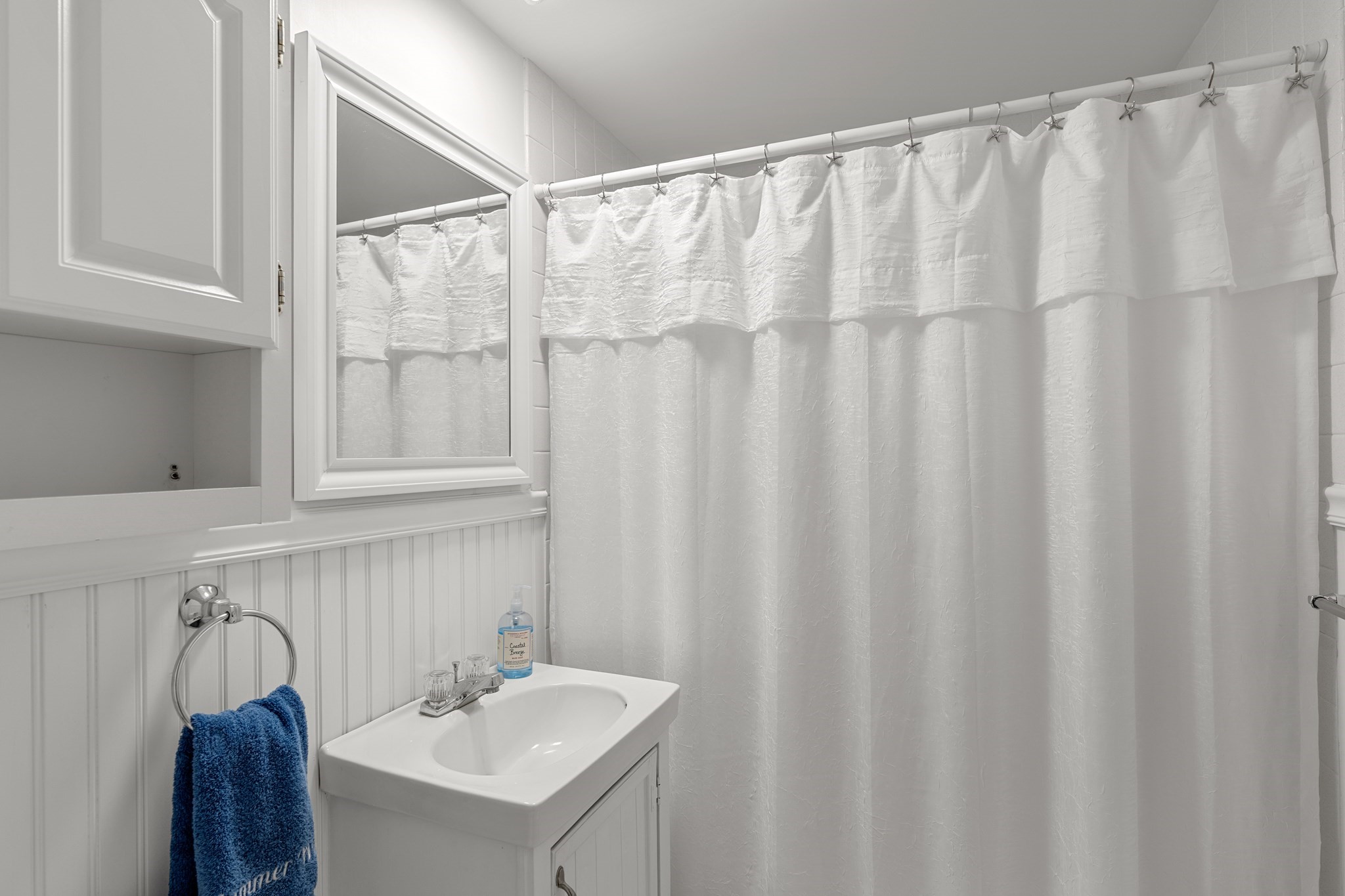 White bathroom with a pedestal-style sink, blue towel, mirrored cabinet, and star-hooked shower curtain.
