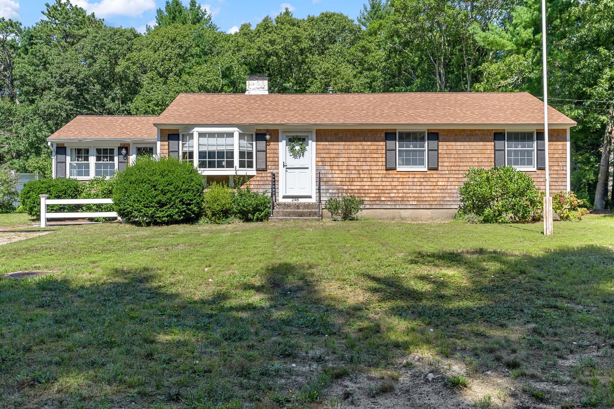 Cedar-shingle single-story house with a white front door, shrubs, and a broad lawn.
