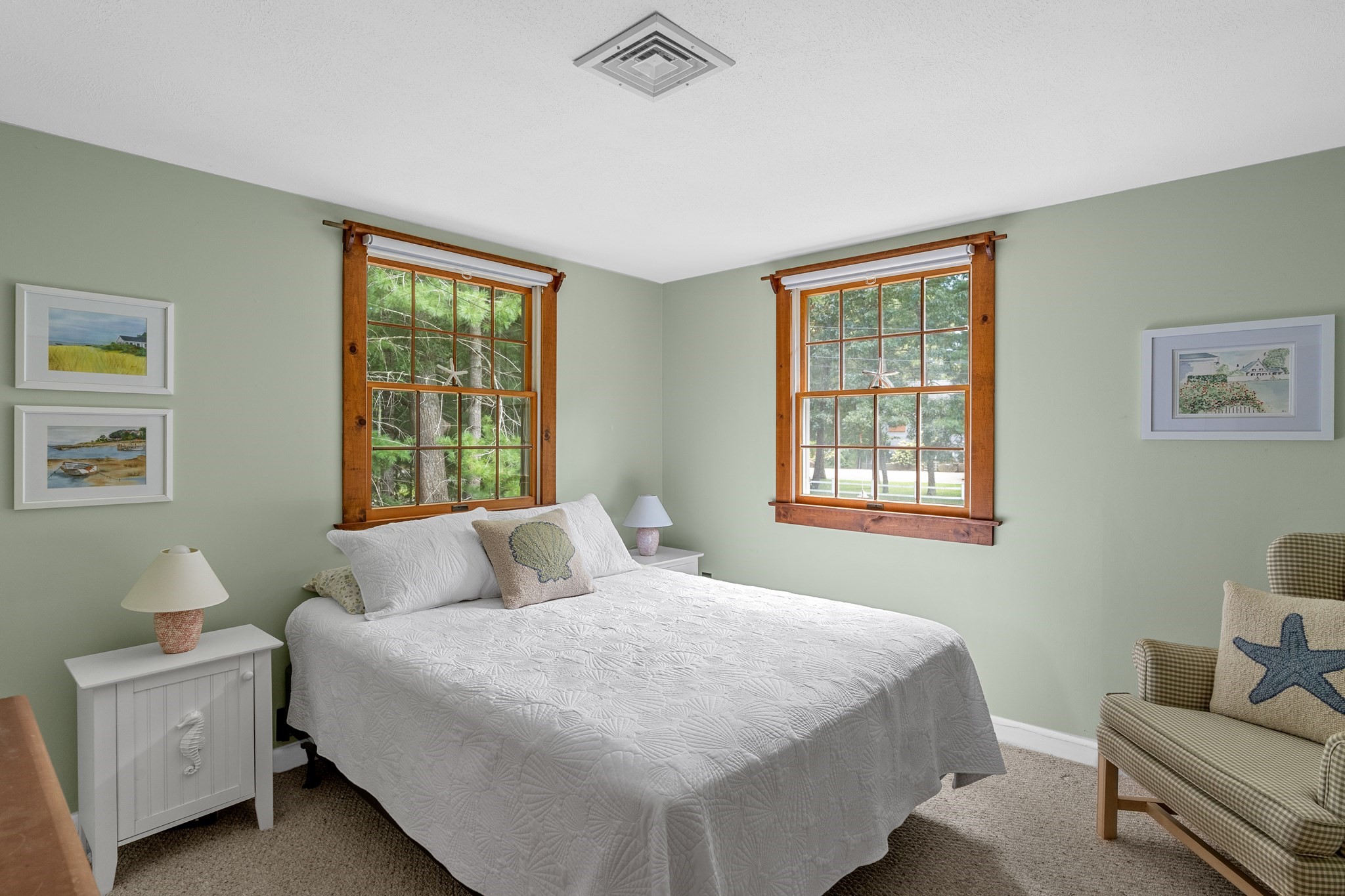 Bedroom with a white quilted bed, sage-green walls, wood-trimmed windows, and a checked armchair.