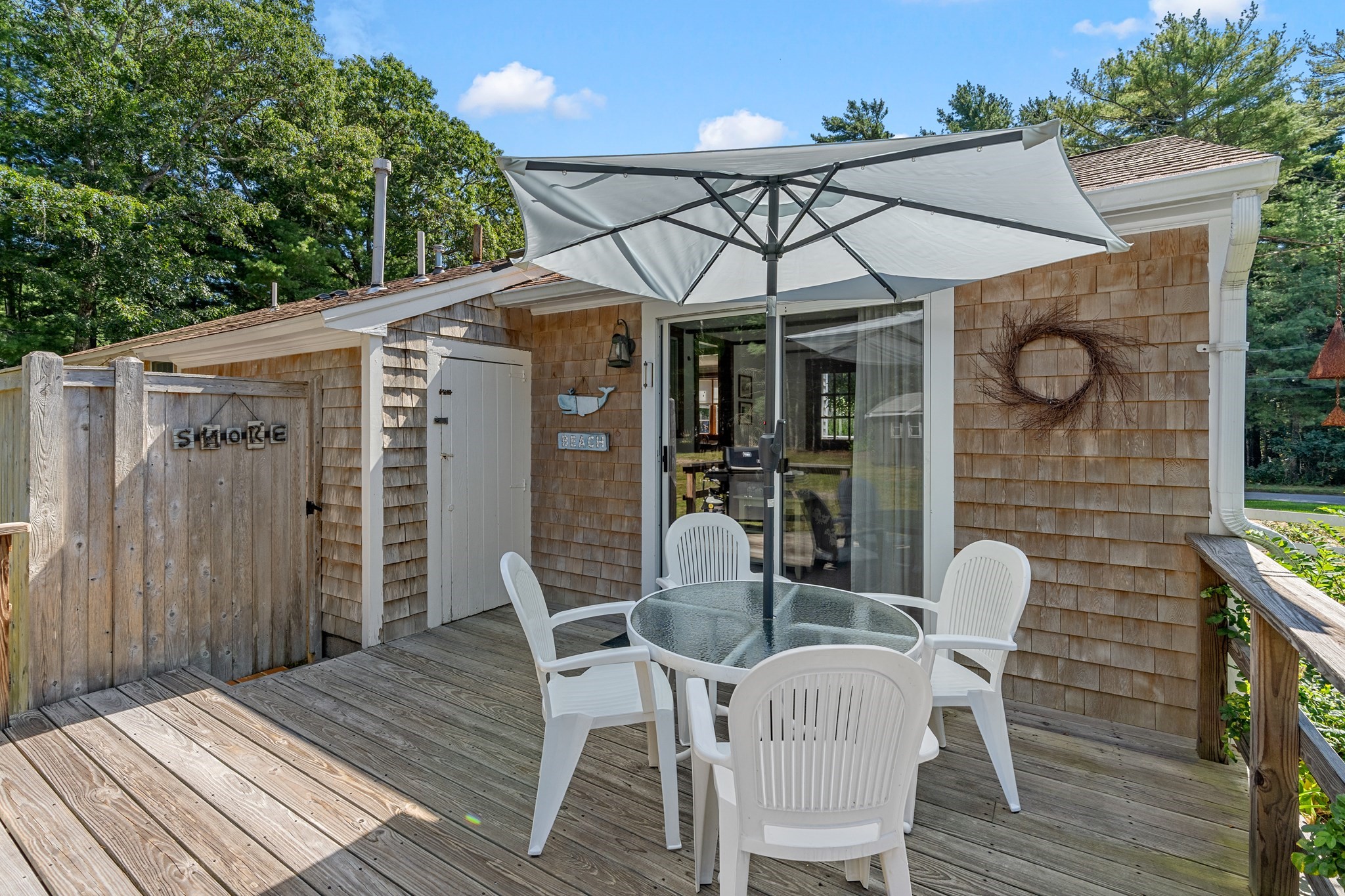 Wooden deck with white patio chairs, a glass-top table, and a large umbrella beside a shingle-sided home.