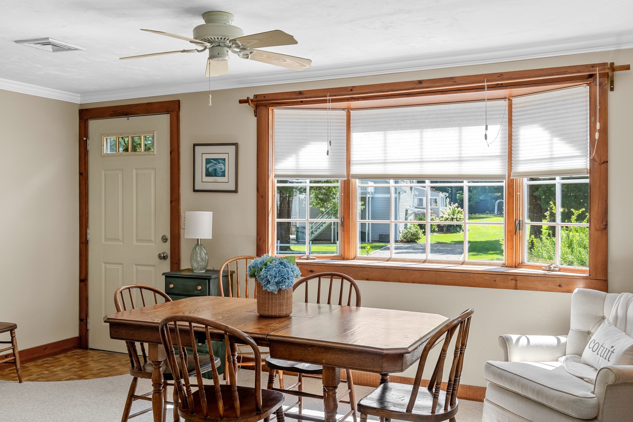 Dining area with a wooden table, blue hydrangeas, and large windows overlooking a green yard.