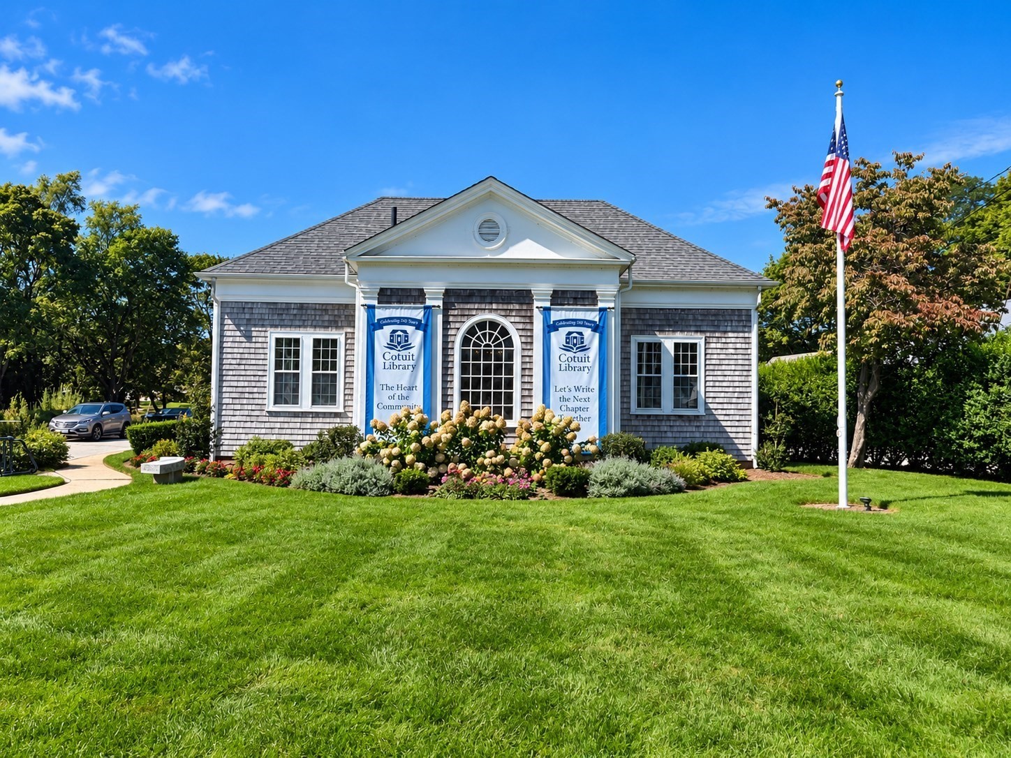 Cotuit Library with blue anniversary banners, a landscaped garden, and an American flag.