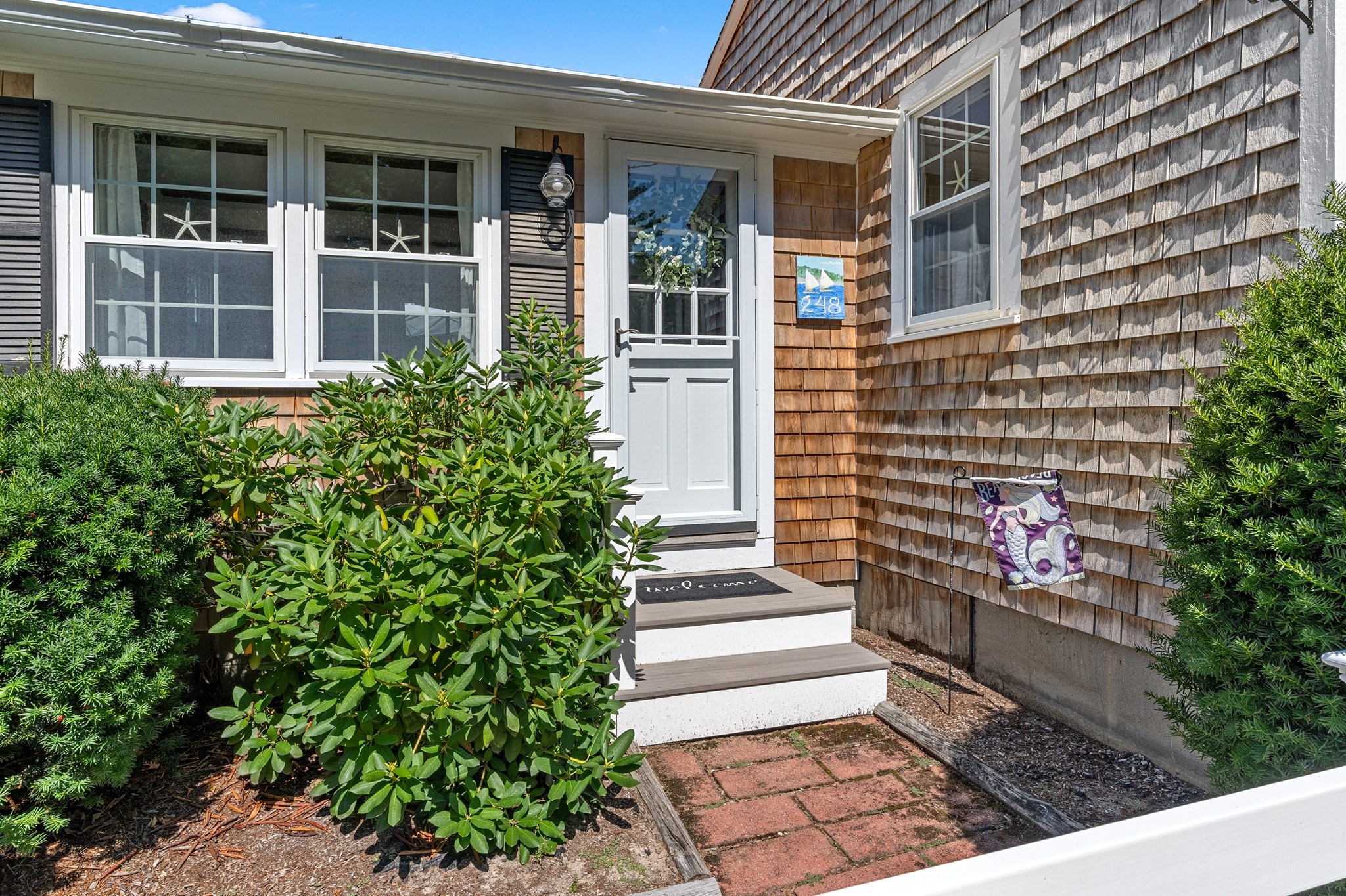 Home entrance with shingle siding, white door, brick path, and leafy shrubs.