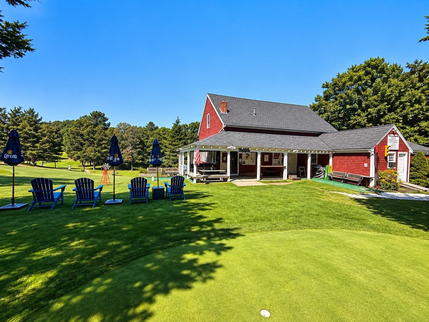 Red-shingled building with a covered porch beside a golf course and lawn seating.