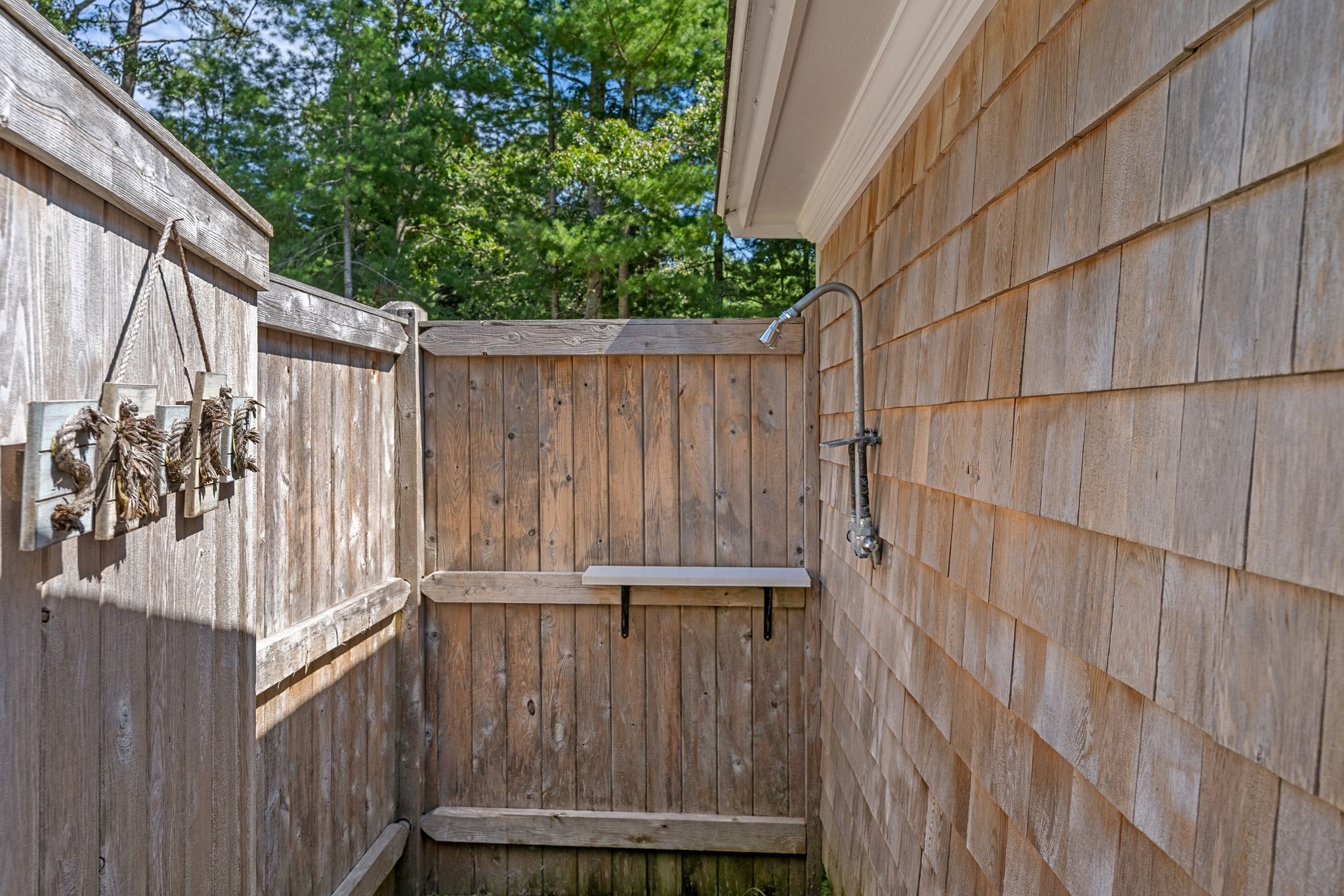 Outdoor shower enclosed by weathered wooden fencing beside cedar-shingled siding, with a small wall-mounted bench.