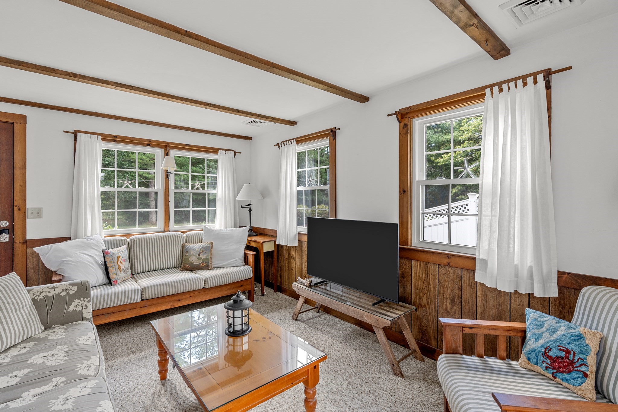 Living room with striped seating, exposed ceiling beams, wood paneling, and bright curtained windows.