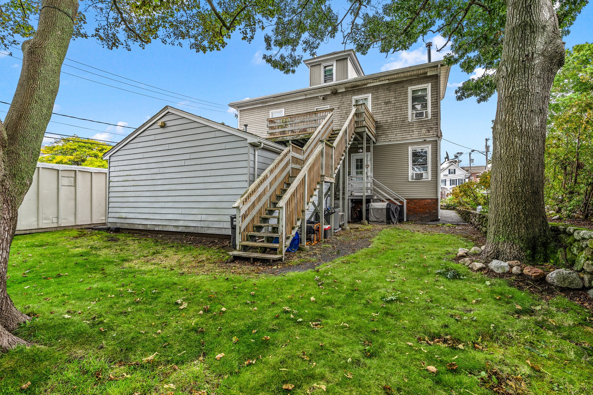 Rear of a gray house with elevated wooden decks and stairs beside a detached garage and tree-lined lawn.