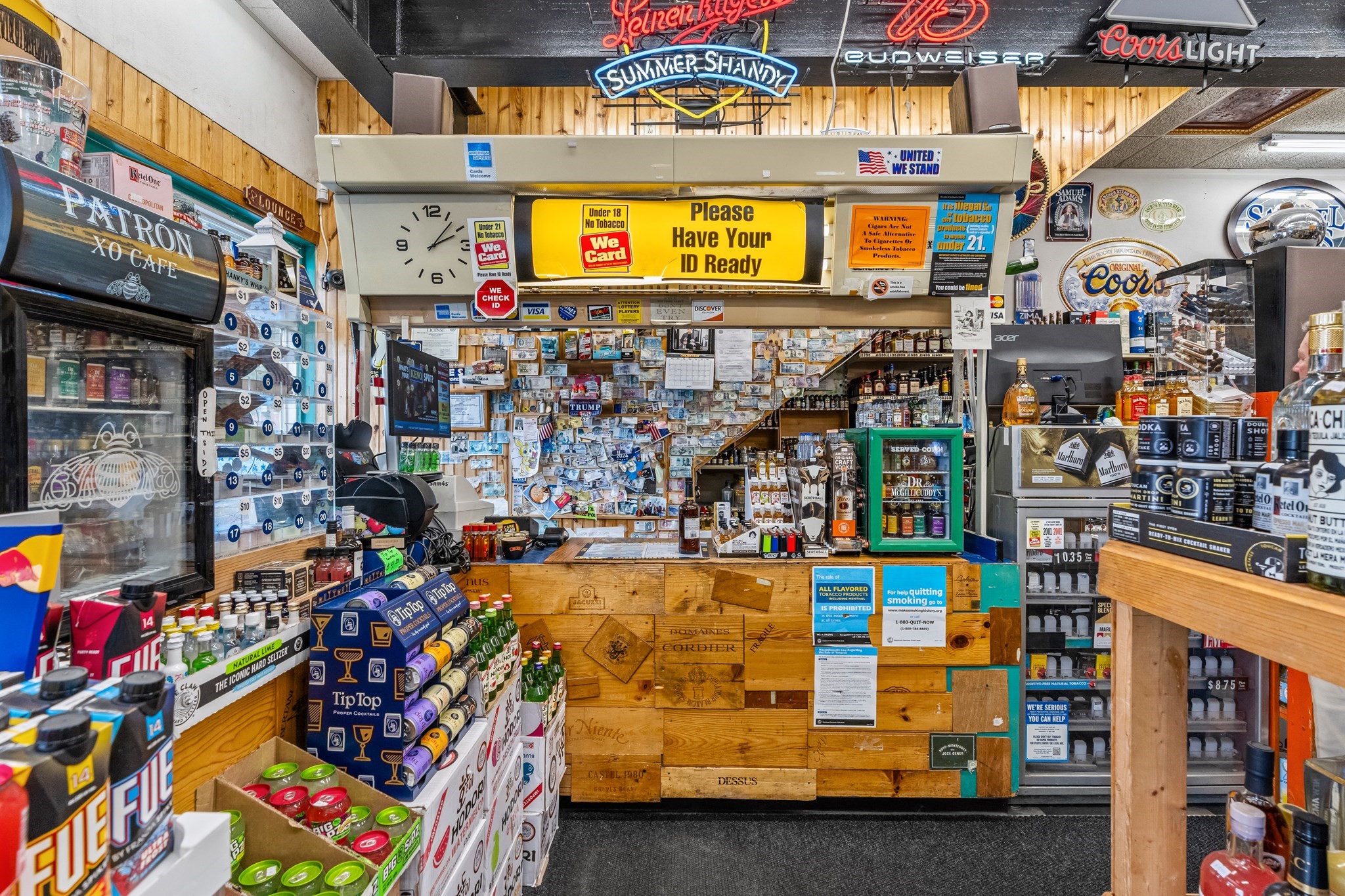 Convenience and liquor store interior with a wooden counter, stocked coolers, and a prominent ID reminder sign.