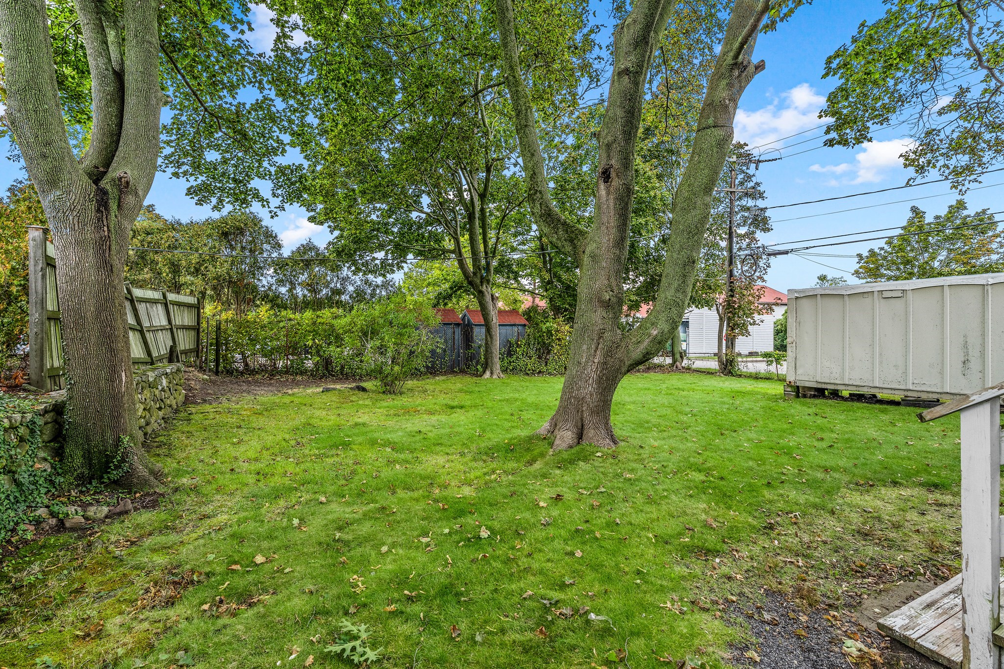 Grassy backyard with mature trees, a stone wall, wooden fence, and storage shed.