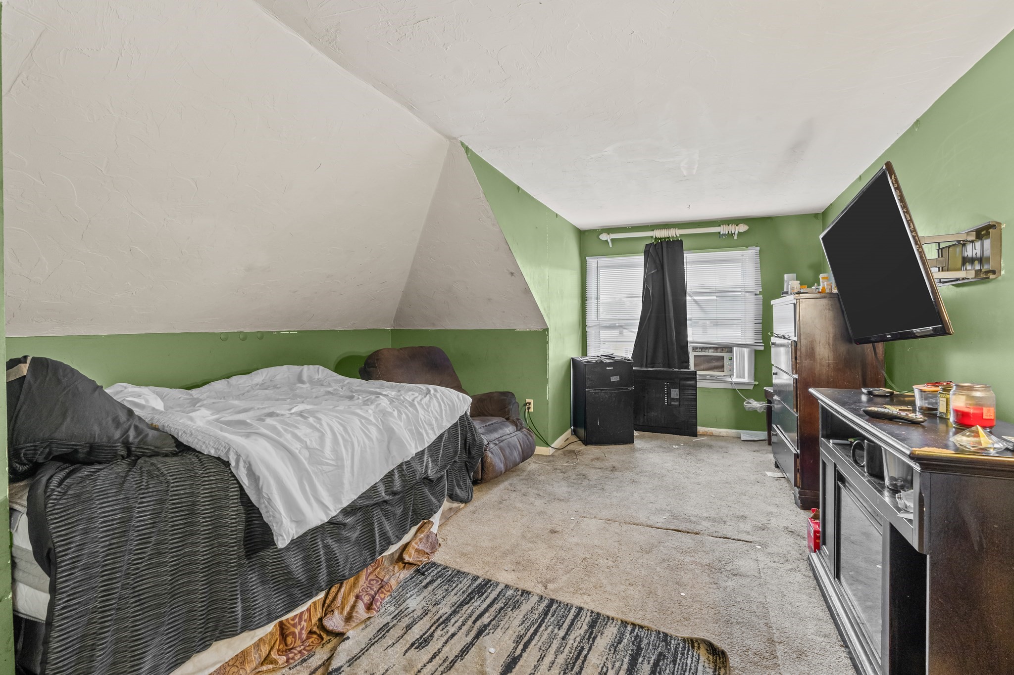 Attic bedroom with green walls, a sloped ceiling, rumpled bed, and a large wall-mounted television.