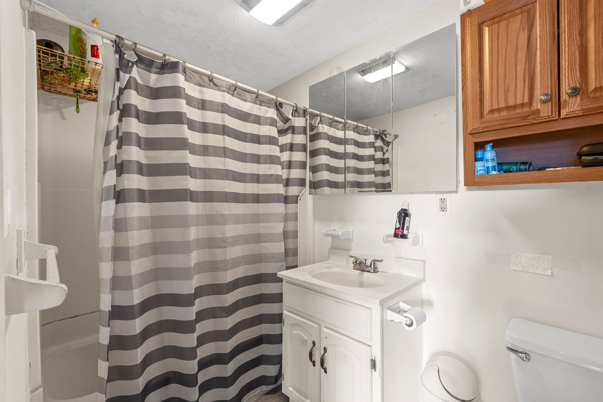 Bathroom with a striped shower curtain, mirrored cabinet, white vanity, and wooden wall cabinet.