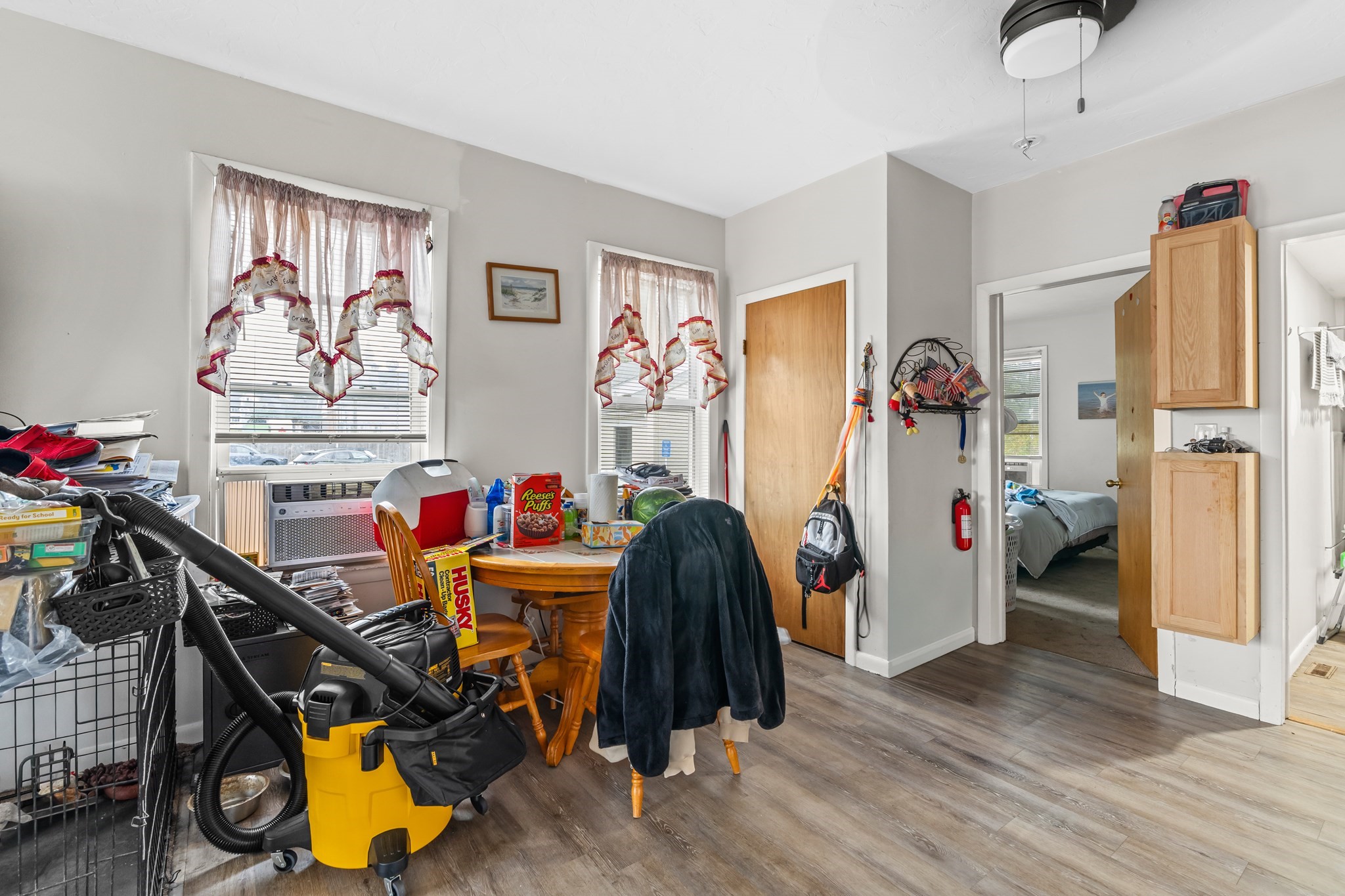 Cluttered dining area with a round wooden table, curtained windows, and a bedroom visible through the doorway.