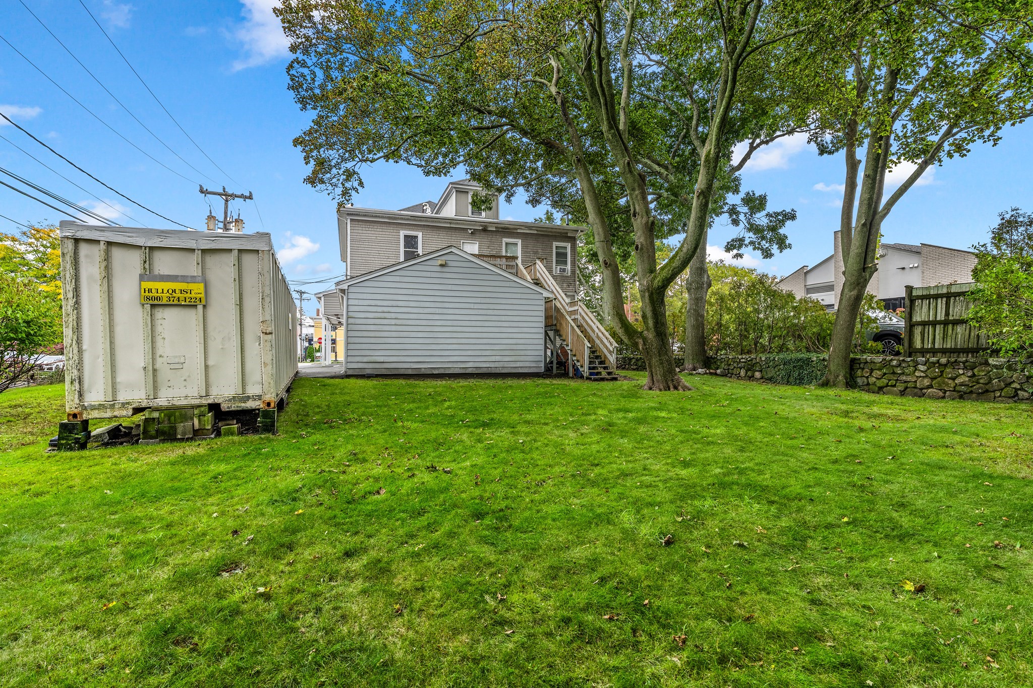 Rear of a gray multi-story property with an outdoor staircase, broad lawn, and mature trees.