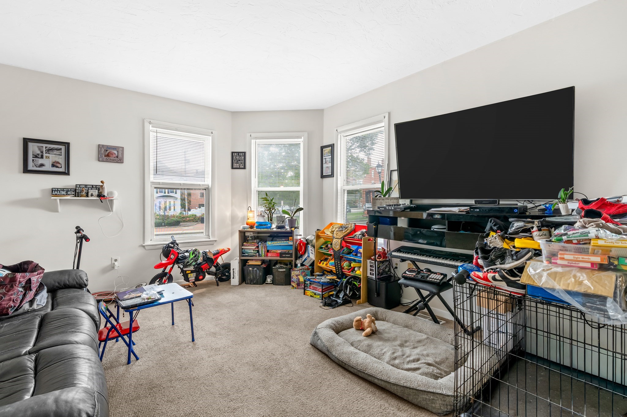 Carpeted living room with three windows, a large television, children's toys, and a dog bed beside a pet pen.