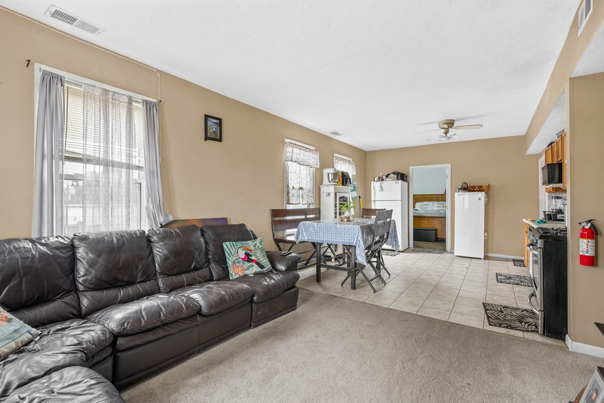 Open-plan living and dining area with a dark leather sectional, tiled floor, and two refrigerators.