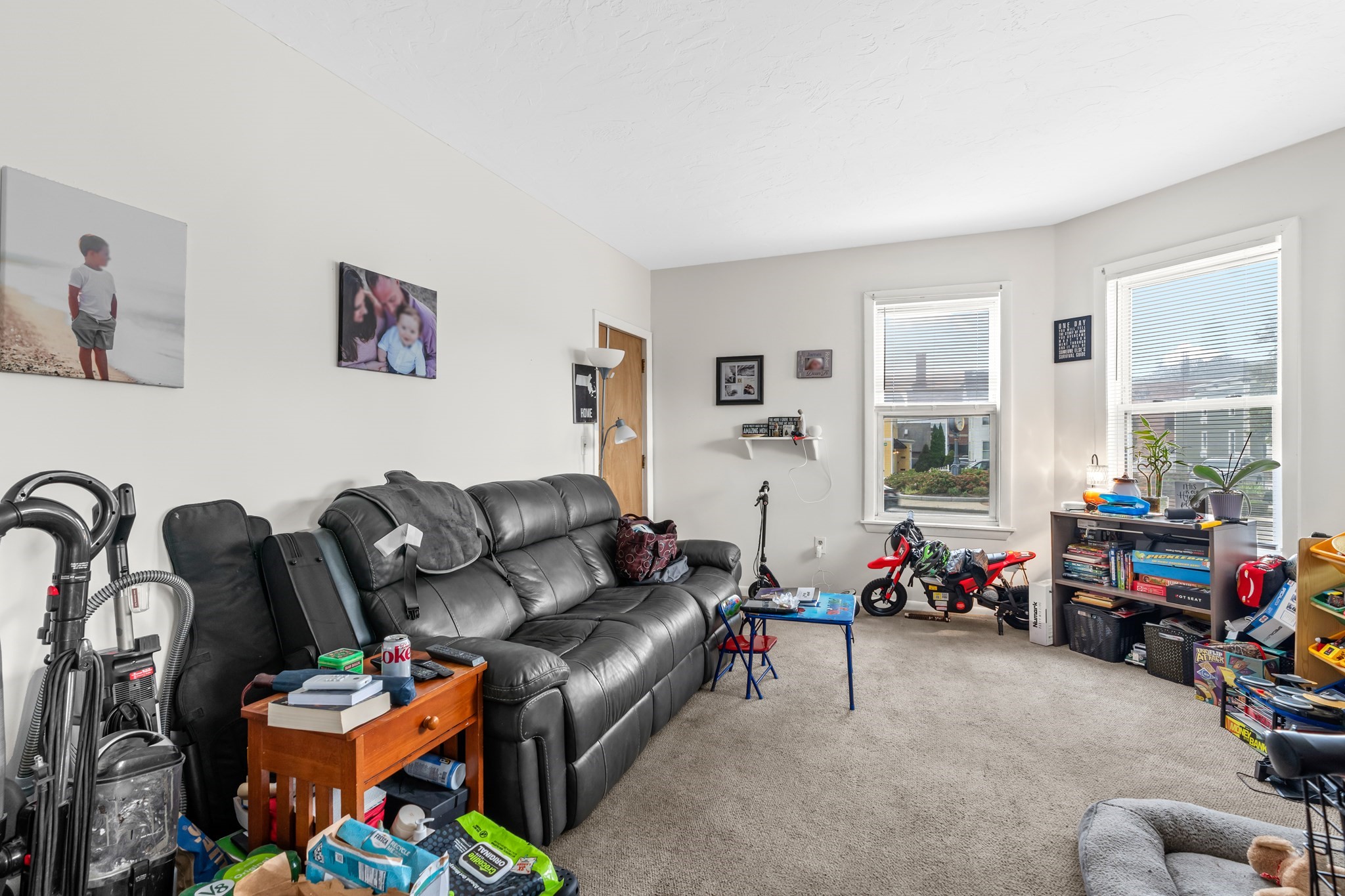 Furnished living room with a black leather sectional, beige carpet, and toys by the windows.
