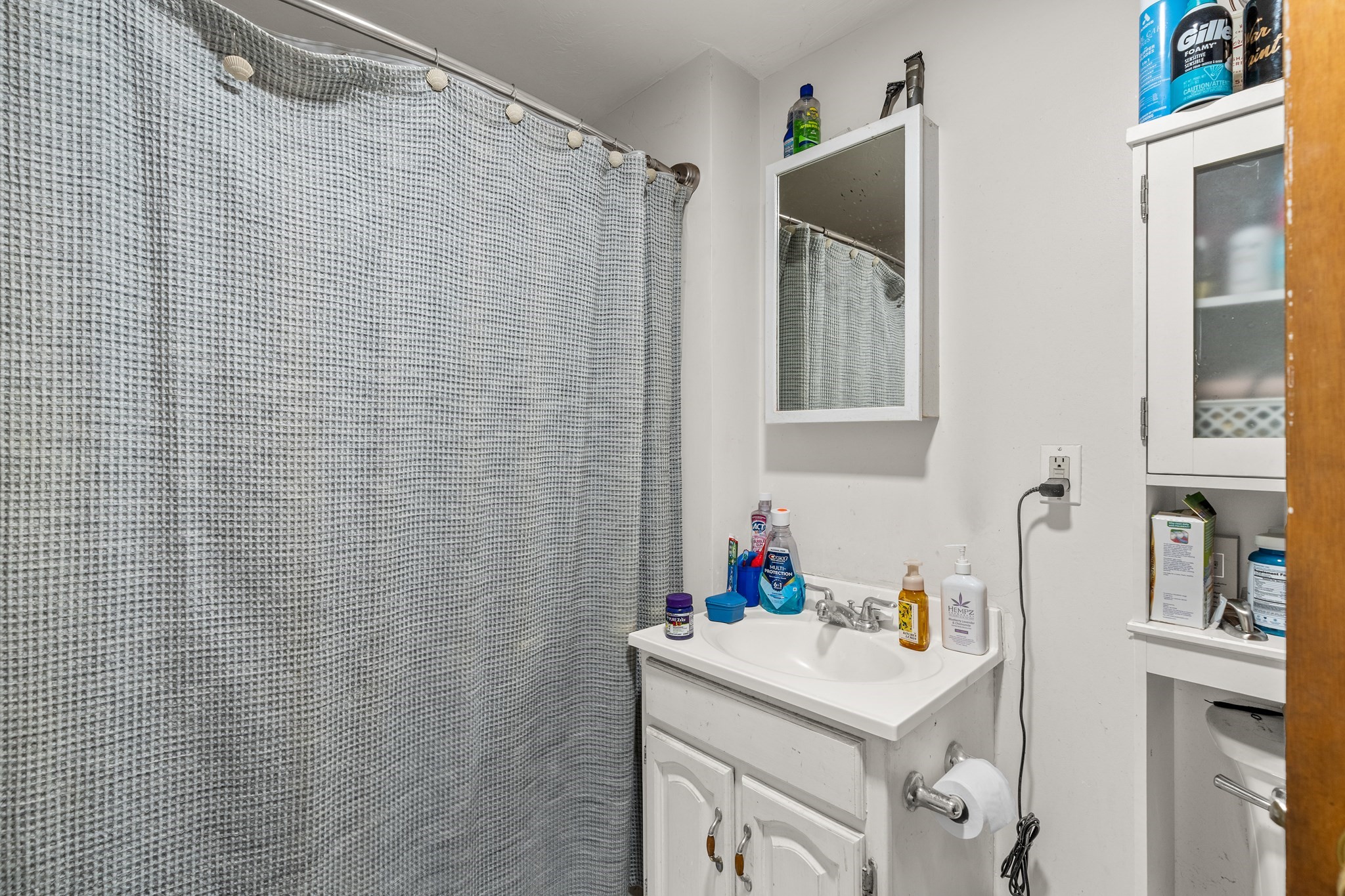 Bathroom with a gray patterned shower curtain, white vanity sink, wall mirror, and mirrored storage cabinet.