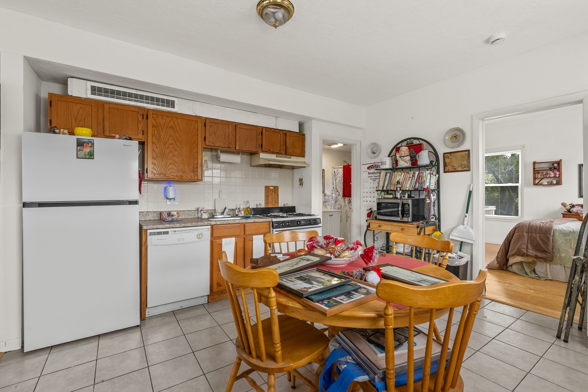 Kitchen and dining area with wood cabinets, a crowded table, and a bedroom visible through the doorway.