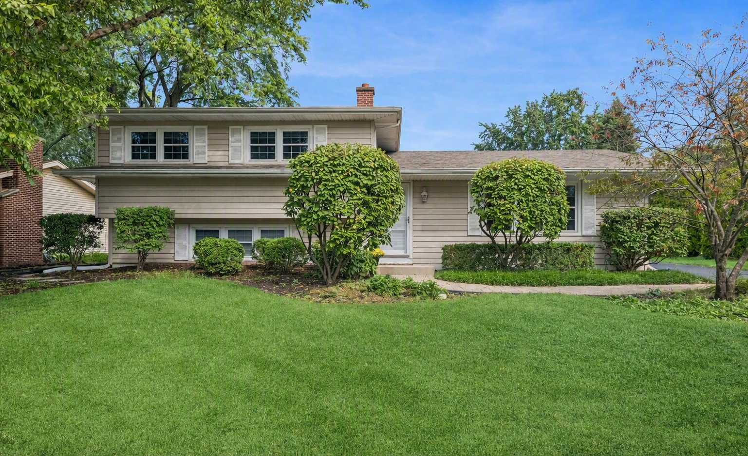 Beige split-level house with white shutters, a brick chimney, and trimmed shrubs behind a broad lawn.