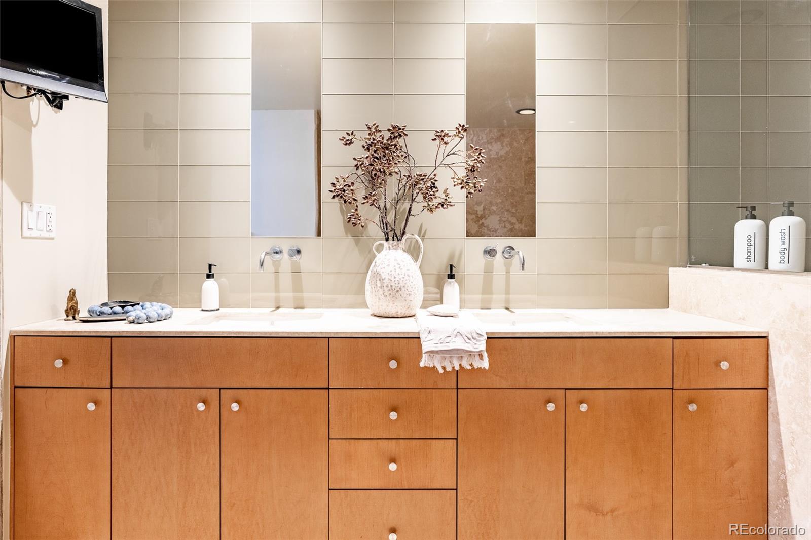 Bathroom vanity with double sinks, warm wood cabinets, tiled wall, and a vase of branches.